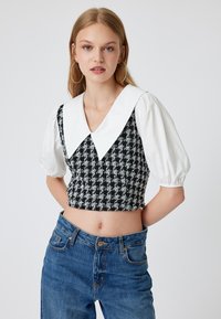 Mustekkon ja valkoisen houndstooth-kuvioinen crop-top, jossa on suuri valkoinen kaulus ja puffatut lyhyet hihat, yhdistettynä korkeavyötäröisiin sinisiin farkkuihin.