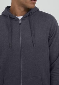 Tamsiai pilkas zip-up džemperis, pagamintas iš minkštos medžiagos. Jame yra kengūros kišenė, raišteliai ir mažas prekės ženklo logotipas ant užtrauktuko.