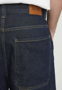 Jeans en denim avec un lavage bleu foncé, présentant des coutures jaunes, une poche arrière et une étiquette en cuir marron embossée avec "BZB."