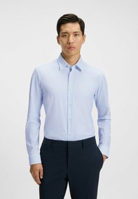 Vaaleansininen pitkähihainen paita hienovaraisella kuviolla, napitettava kaulus ja nappisuljenta edessä. Yhdistetty tummaan housuun, joka näyttää mukautetulta.