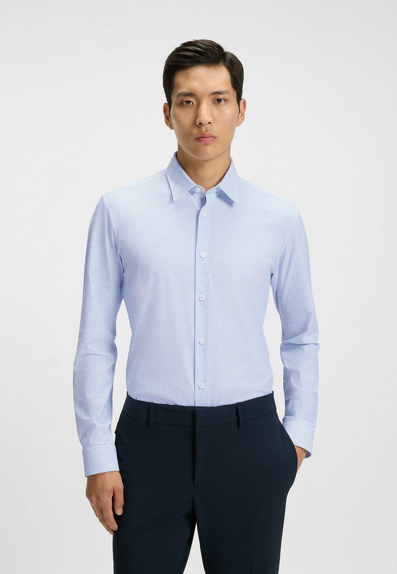 Vaaleansininen pitkähihainen paita hienovaraisella kuviolla, napitettava kaulus ja nappisuljenta edessä. Yhdistetty tummaan housuun, joka näyttää mukautetulta.