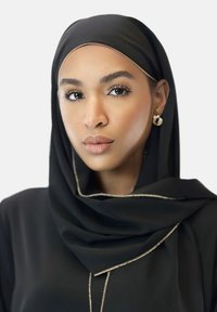 Schwarzer Hijab mit goldenen Akzenten, glattes Stoff, umrahmt das Gesicht; Modell mit neutralem Make-up, sichtbaren Ohrringen, sanftes Licht auf der Haut.