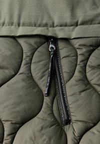 Tissu olive vert matelassé avec des motifs arrondis. Présente une fermeture éclair noire avec une languette en textile et une couche supérieure lisse.