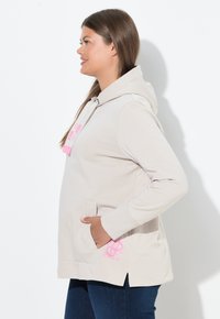 Hoodie beige chiaro con tasca frontale, dettagli rosa, lacci regolabili al collo e spacchi laterali. Texture liscia e design casual.