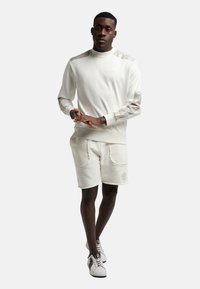Cremiger Pullover mit gemusterten Ärmeln, kombiniert mit cremigen Shorts mit zwei Fronttaschen. Das Model trägt weiße Turnschuhe.