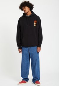 Zwarte katoenen hoodie met een grafisch logo op de borst, gecombineerd met losse, blauwe denim broek en rode instappers.