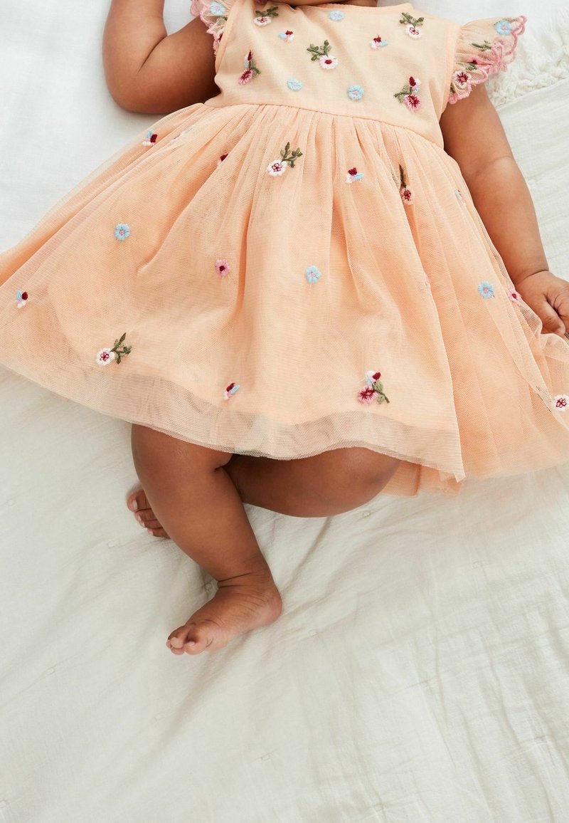 Baby in einem Pfirsichkleid mit gestickten Blumen, liegt auf einer weißen Stoffoberfläche und zeigt Beine und einen Teil des Arms.