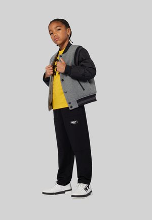 Grijze en zwarte bomberjack over een gele shirt, zwarte cargo-broek en witte sneakers met zwarte accenten. Logo's op jack en broek.