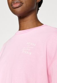 Jasnoróżowa bawełniana bluza z okrągłym dekoltem, z białym napisem "ADIDAS Three Stripes STUDIO" na lewej piersi.