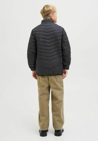 Jachetă puf de culoare neagră cu model în zig-zag, guler înalt și detalii cusute, purtată peste pantaloni cargo bej și pantofi negri.