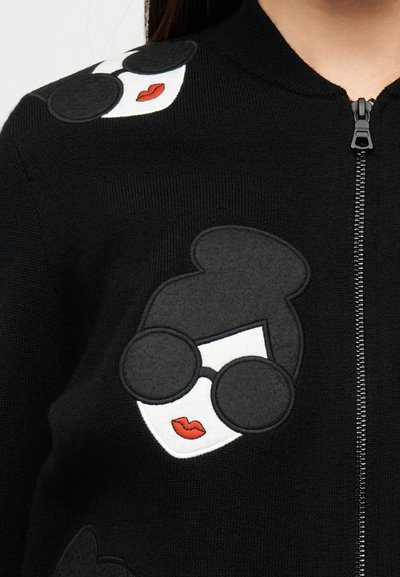 Crni pleteni džemper na patentni zatvarač s bijelim i crnim izvezenim licima s naočalama za sunce i crvenim usnama na prednjoj strani. Glatka tekstura.