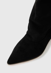 Black suede ankle boot with a pointed toe, smooth texture, and fitted shaft. Seam detail at the base, set against a light background.
