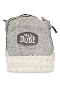 Zapato slip-on de tela gris con un acabado texturizado, que presenta una suela moteada blanca y un parche que dice "HEY DUDE" en la parte posterior.