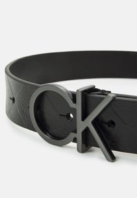 Cintura in pelle nera con superficie testurizzata e fibbia metallica circolare con un design sovrapposto "CK" in nero opaco.