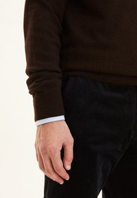 Brauner Pullover mit gerippten Bündchen, kombiniert mit dunkler Cordswehose. Der kragen eines hellblauen Hemdes ist unter dem Pullover sichtbar.