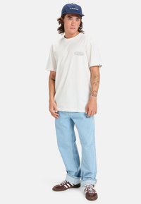 Hvid kortærmet t-shirt med lille blåt logo. Lyseblå denimjeans med opbrudte cuff. Mørkebrune sneakers med hvide striber og snørebånd. Marineblå cap.