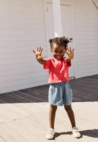 Barn i en rød kortærmet T-shirt og lyseblå denim shorts, smilende med hænderne løftet. Bærer hvide sneakers med farverige remme.