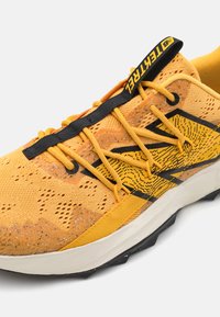 Shoe de course en mesh jaune respirant avec des accents noirs, un motif texturé et des points en relief ; dispose de lacets noirs et d'une semelle blanche.