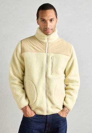 Veste en polaire couleur crème avec col montant, panneau d'épaule beige, poche zippée à l'avant et coutures latérales courbées. Surface douce et texturée.