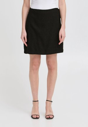 Vrouw draagt een zwarte knielange rok, een wit topje en zwarte hoge sandalen met enkelbandje, staand op een witte achtergrond.