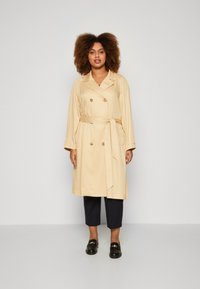 Trench beige con botones de doble botonadura, cinturón, bolsillos laterales y mangas largas, combinado con pantalones negros cortos y zapatos negros.