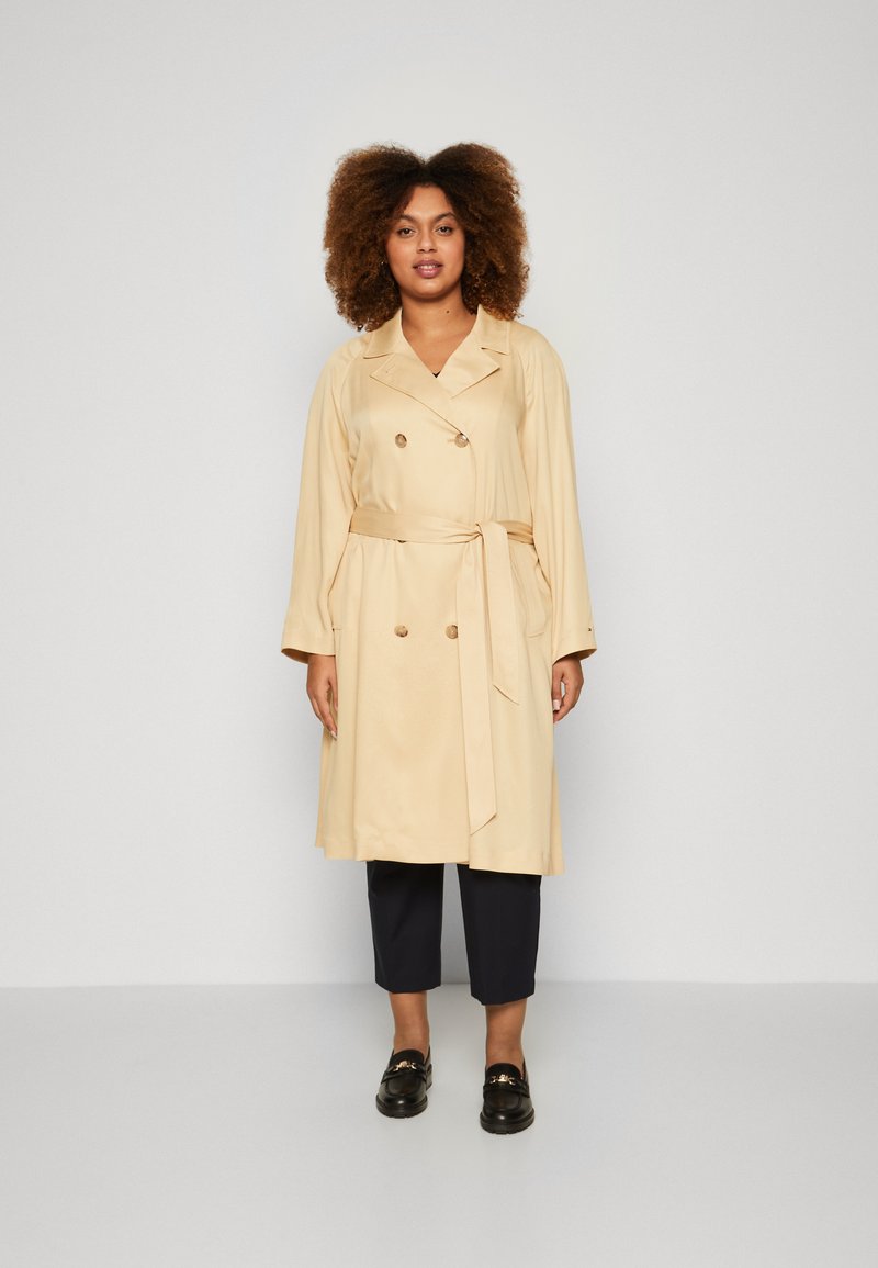 Trench beige con botones de doble botonadura, cinturón, bolsillos laterales y mangas largas, combinado con pantalones negros cortos y zapatos negros.