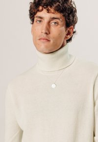Cremfarbener Rollkragenpullover aus weichem, strukturiertem Strick. Verfügt über eine silberne Anhängerkette mit einem runden, glänzenden Detail.