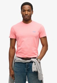 Roze T-shirt met korte mouwen van katoen, voorzien van een klein blauw logo op de borst. Draag het met donkere spijkerbroeken en een grijze trui om de taille geknoopt.