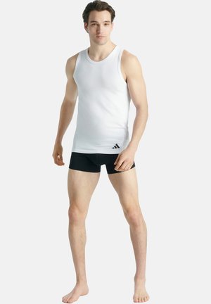 Weißes ärmelloses Tanktop mit rundem Ausschnitt, getragen über schwarzen Shorts. Verfügt über ein dezentes Branding auf der unteren linken Seite.