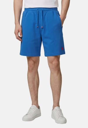 Man draagt felblauwe shorts met koordje en zijzakken en witte sneakers, staand met ontspannen handen naast het lichaam.