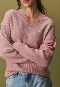 Rosa geripptes Sweatshirt mit V-Ausschnitt und weiten Ärmeln, kombiniert mit hellen Jeans aus Denim mit Verzierungen. Strukturiertes Strickgewebe.