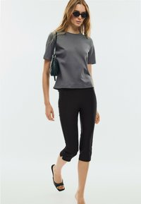 Jeune femme portant des lunettes de soleil foncées, un t-shirt gris à manches courtes, un pantalon court noir, des sandales noires à enfiler, et un sac à bandoulière noir.