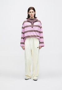 Νεαρή γυναίκα στέκεται, φορώντας ένα ροζ και καφέ ριγέ μπλουζάκι με μακριά μανίκια και χαλαρούς λευκούς παντελόνες με μαύρα αθλητικά παπούτσια.