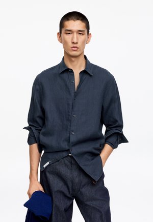 Jeune homme aux cheveux courts portant une chemise bleu foncé boutonnée et un pantalon sombre, tenant un vêtement bleu plié dans sa main gauche.