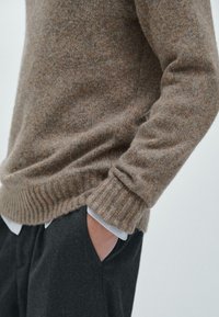 Strickpullover in einem braunen und beigen Mélange, mit gerippten Ärmelabschlüssen und Saum, kombiniert mit dunklen Hosen. Hände in den Taschen.