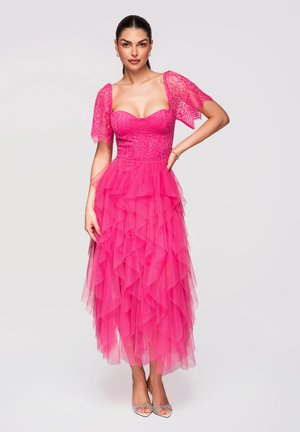 Kobieta stoi z jedną ręką na biodrze, ubrana w jasnoróżową koronkową sukienkę z marszczoną tiulową spódnicą i przezroczystymi bufkami.