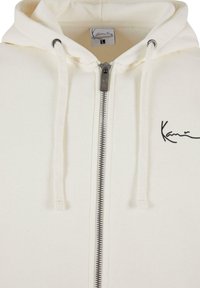 Hvit zip-up hettegenser med snor, med et brodert svart logo. Mykt teksturert stoff og sølvfarget glidelås.