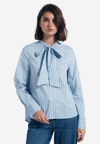 Camicia a righe blu chiaro con colletto a farfallino, maniche lunghe e chiusura con bottoni. Realizzata in tessuto liscio con righe verticali di un blu più scuro.