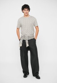 Hombre con cabello rizado que lleva una camisa de manga corta estampada, un suéter a juego atado a la cintura, pantalones cargo negros anchos y zapatos negros, de pie con las manos en los bolsillos.