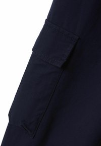 Navyblauwe stof met een gladde textuur, voorzien van een patchzak; de stiksels langs de randen zijn zichtbaar en verhogen de functionaliteit van het kledingstuk.