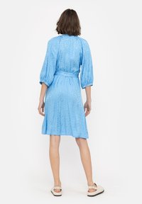 Niebieska sukienka z gładkiej tkaniny, z rękawami do łokcia, wcięciem w talii i rozkloszowanym dołem. Posiada detal z wiązaniem z tyłu. Noszone płaskie sandały.