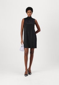 Svart ärmlös klänning med hög halsringning, tillverkad av glittrande material. Kombinerad med en liten lavendelfärgad handväska och svarta spetsiga klackar.