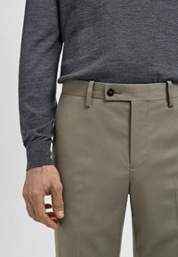 Mann trägt ein graues Langarmhemd, das in beige Hose mit Knopf und Gürtelschlaufen gesteckt ist, Hand entspannt an der Seite.