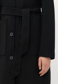 Cappotto di lana nero con cintura in vita annodata, due tasche frontali, chiusura con bottoni e una mano poggiata accanto alla manica del cappotto.
