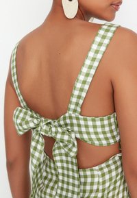 Grün-weißer Gingham-Kleid mit tiefem Rücken, breiten Trägern und einem großen Schleifendetail an der Taille, das ein strukturiertes Gewebe zeigt.