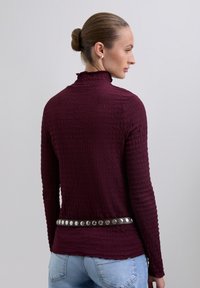 Burgundy long-sleeve top with a high ruffled collar and textured fabric, featuring a silver studded belt at the waist, shown from behind.