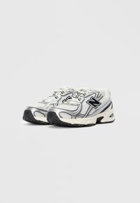 Une paire de baskets de sport blanches et argentées avec des semelles noires et un logo "N" sur les côtés, positionnées de manière angulaire sur un fond blanc uni.