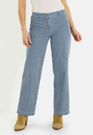 Mujer con pantalones de rayas verticales azules y blancas, top sin mangas blanco y botines de ante marrón claro, de pie con las manos relajadas.