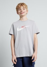 Šviesiai pilkas medvilninis marškinėliai su dideliu baltu Nike swoosh, mažesniu raudonu swoosh ir tekstu "Nike Athletic Dept."