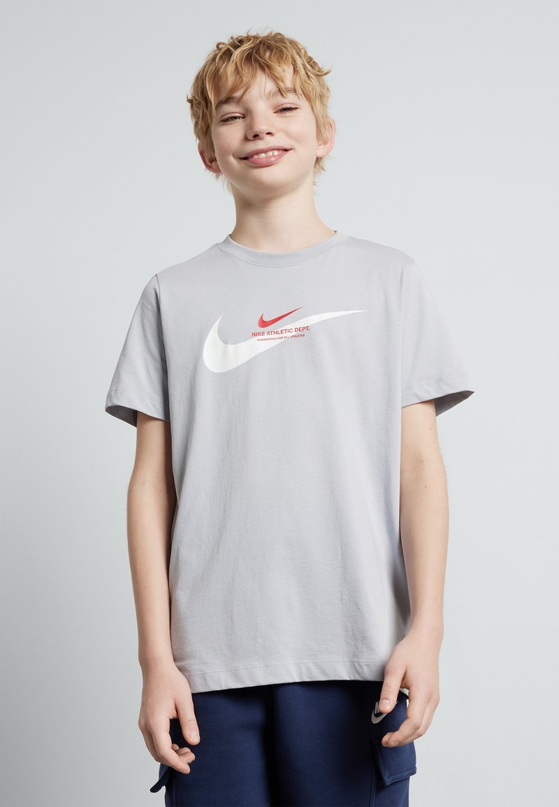 Šviesiai pilkas medvilninis marškinėliai su dideliu baltu Nike swoosh, mažesniu raudonu swoosh ir tekstu "Nike Athletic Dept."